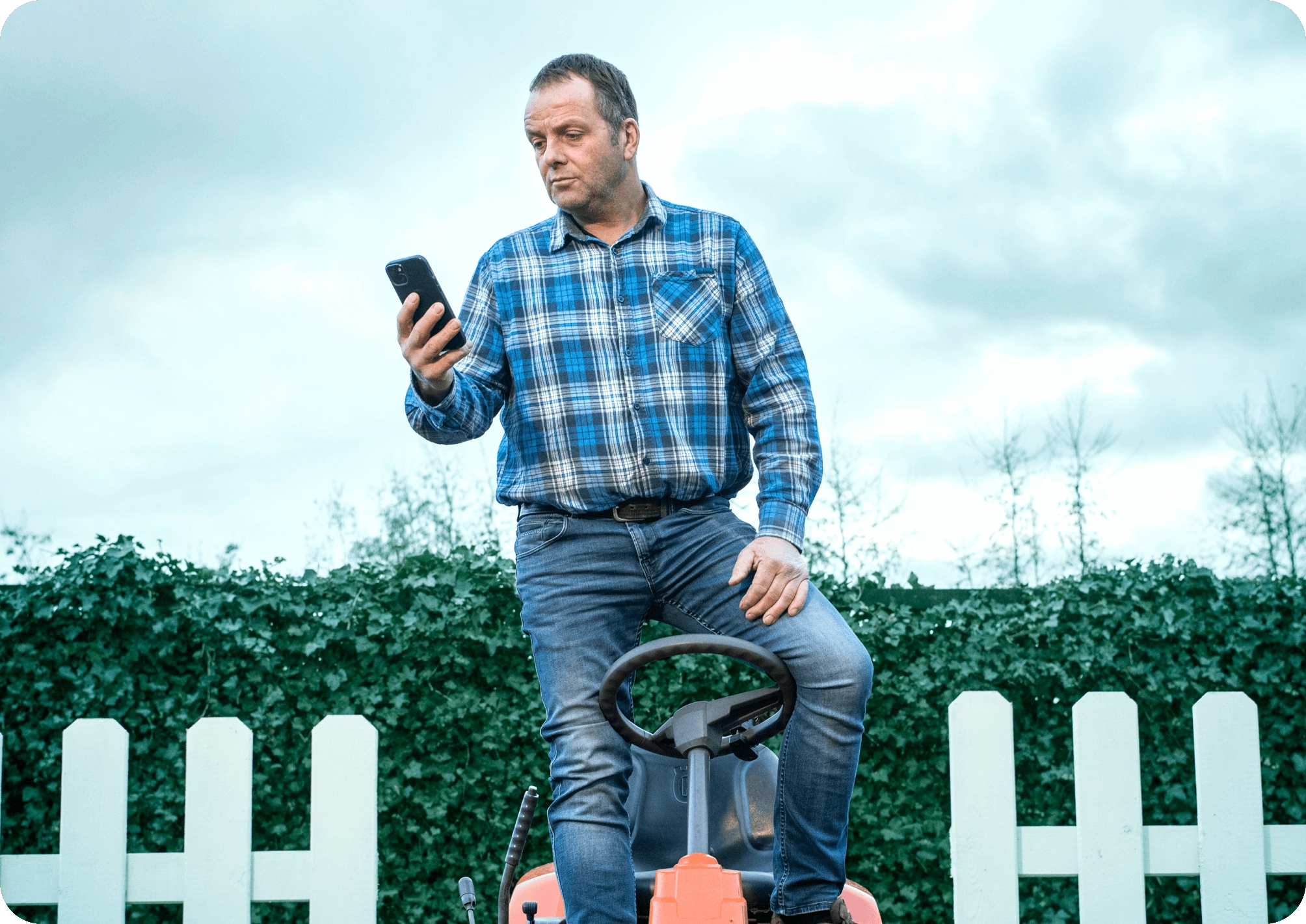 Man standing on lawn tractor outdoors, looking at smartphone under cloudy sky.